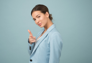 light blue plain background wit a young lady with brown hair standing in front of it. She is wear a blue blazer and is pointing her finger as well as looking at you 