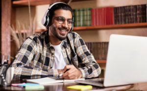 Young man with earphones on sitting at a desk talking to someone onnhis laptop. The young man is smilling and taking notes
