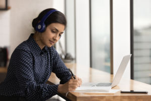 Women wearing beats headphones writting on a document sitting at a desk with a laptop open in front of her