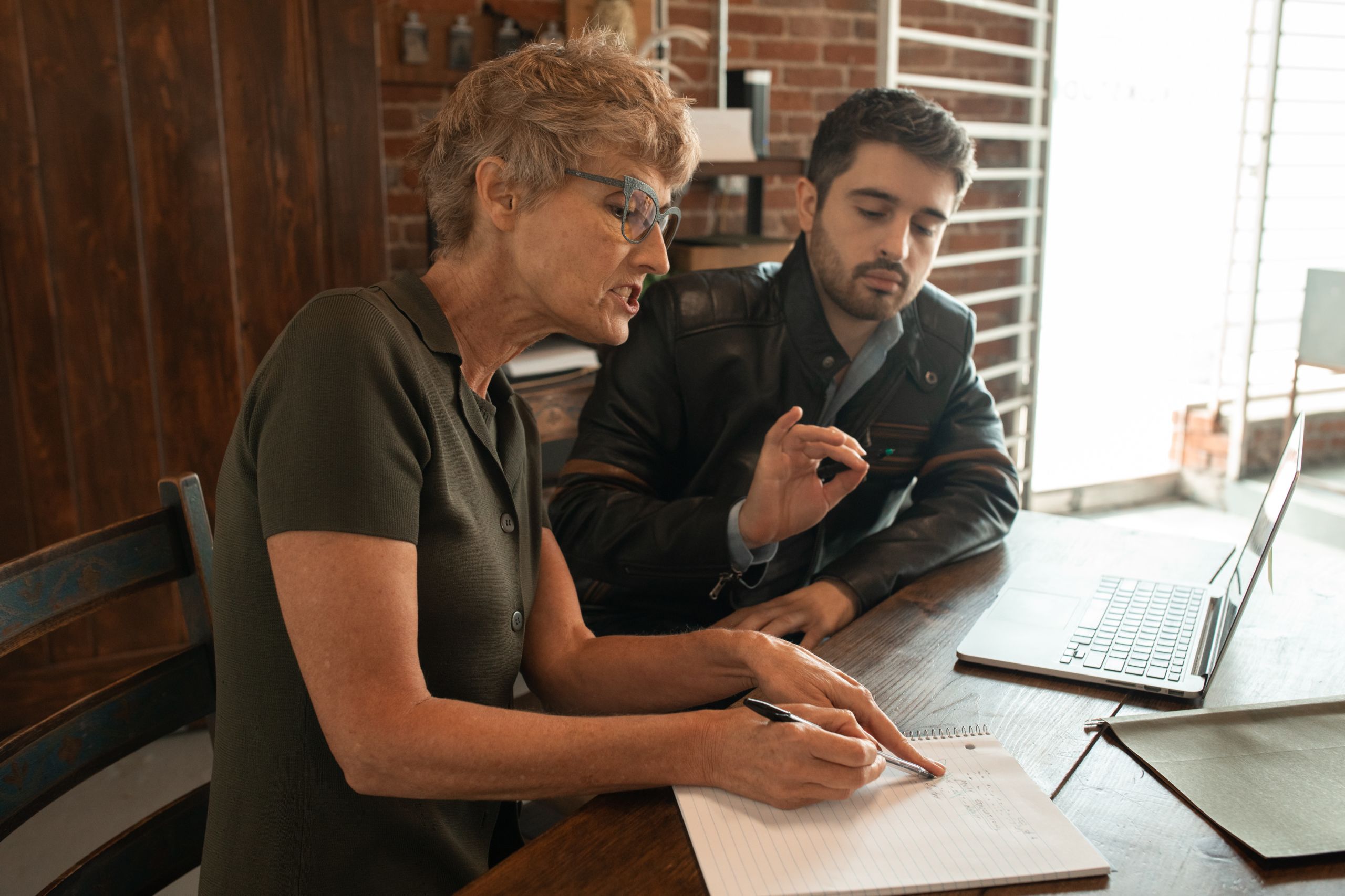 older lady with short hair and glasses talking to a younger guy sitting next to her. She is writting on a peice of paper and the guy next to her is also t.alking and has a laptop in front of him