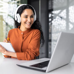 Latin american business woman with curly hair and headphones watching online training course at workplace, woman writing information happy and satisfied with the results of professional development. Latin american business woman with curly hair and headphones watching online training course at workplace, woman writing information happy and satisfied with the results of professional development