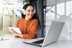 Latin american business woman with curly hair and headphones watching online training course at workplace, woman writing information happy and satisfied with the results of professional development. Latin american business woman with curly hair and headphones watching online training course at workplace, woman writing information happy and satisfied with the results of professional development