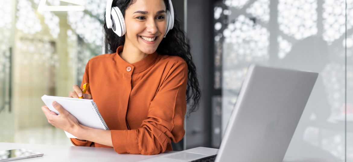 Latin american business woman with curly hair and headphones watching online training course at workplace, woman writing information happy and satisfied with the results of professional development. Latin american business woman with curly hair and headphones watching online training course at workplace, woman writing information happy and satisfied with the results of professional development