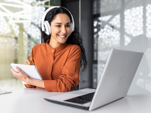 Latin american business woman with curly hair and headphones watching online training course at workplace, woman writing information happy and satisfied with the results of professional development. Latin american business woman with curly hair and headphones watching online training course at workplace, woman writing information happy and satisfied with the results of professional development