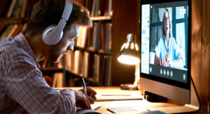 Male student wearing headphones conference video calling, watching webinar, online training class, virtual chat meeting with remote teacher or coach distance learning using computer, taking notes. Male student wearing headphones video conference calling watching webinar.