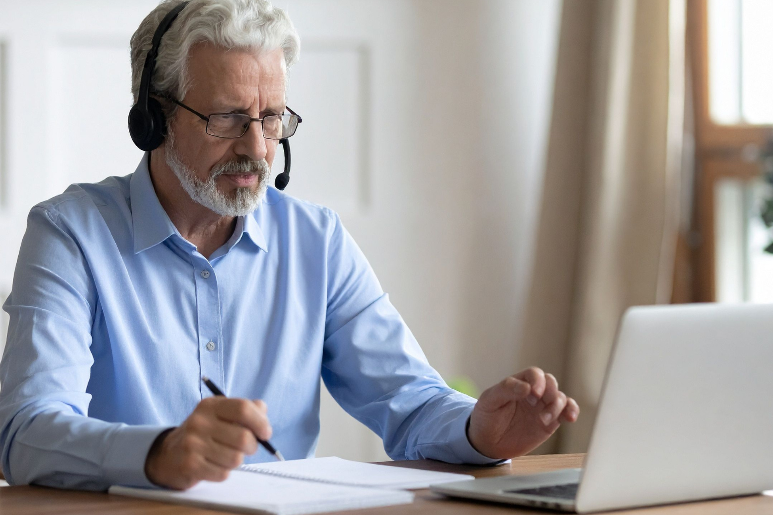 Male professional attending an online language class, wearing a headset and taking notes in front of a laptop